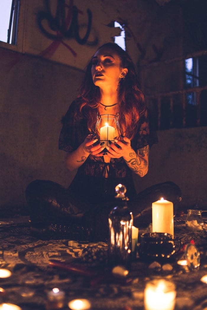 A mysterious woman engages in a ritual, holding a candle in a dimly lit, gothic room.