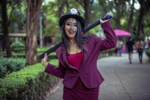 Cosplay performer wearing skull-adorned hat and makeup poses outdoors in a vibrant park setting.