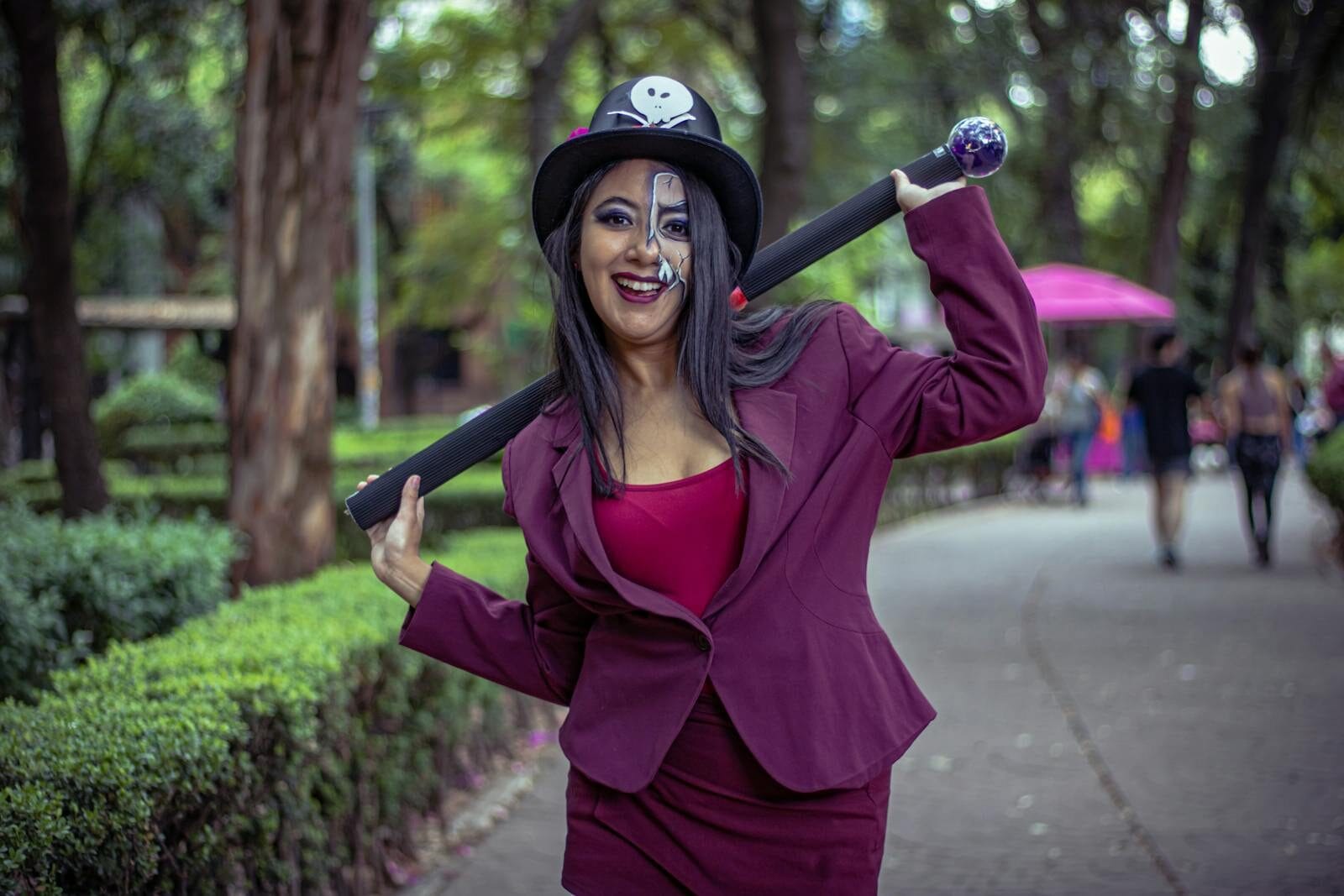 Cosplay performer wearing skull-adorned hat and makeup poses outdoors in a vibrant park setting.