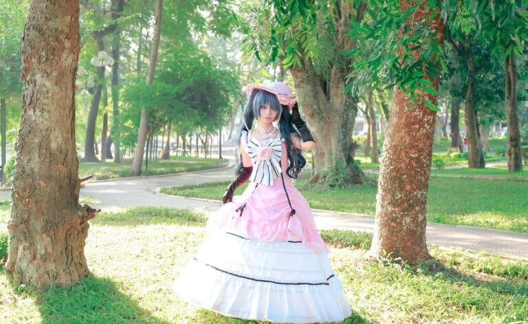A Cosplayer In A Pink Victorian Gown Poses In A Sunny Park With Lush Greenery.