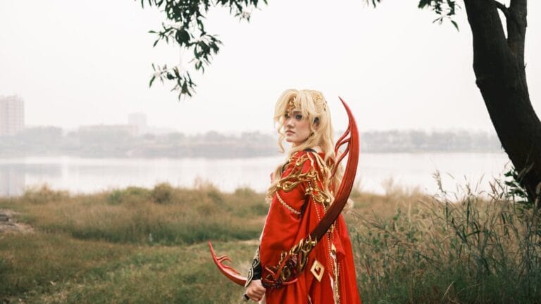 A Person In A Fantasy Cosplay Outfit With A Red Costume Stands By A Lakeside, Holding A Decorative Bow.