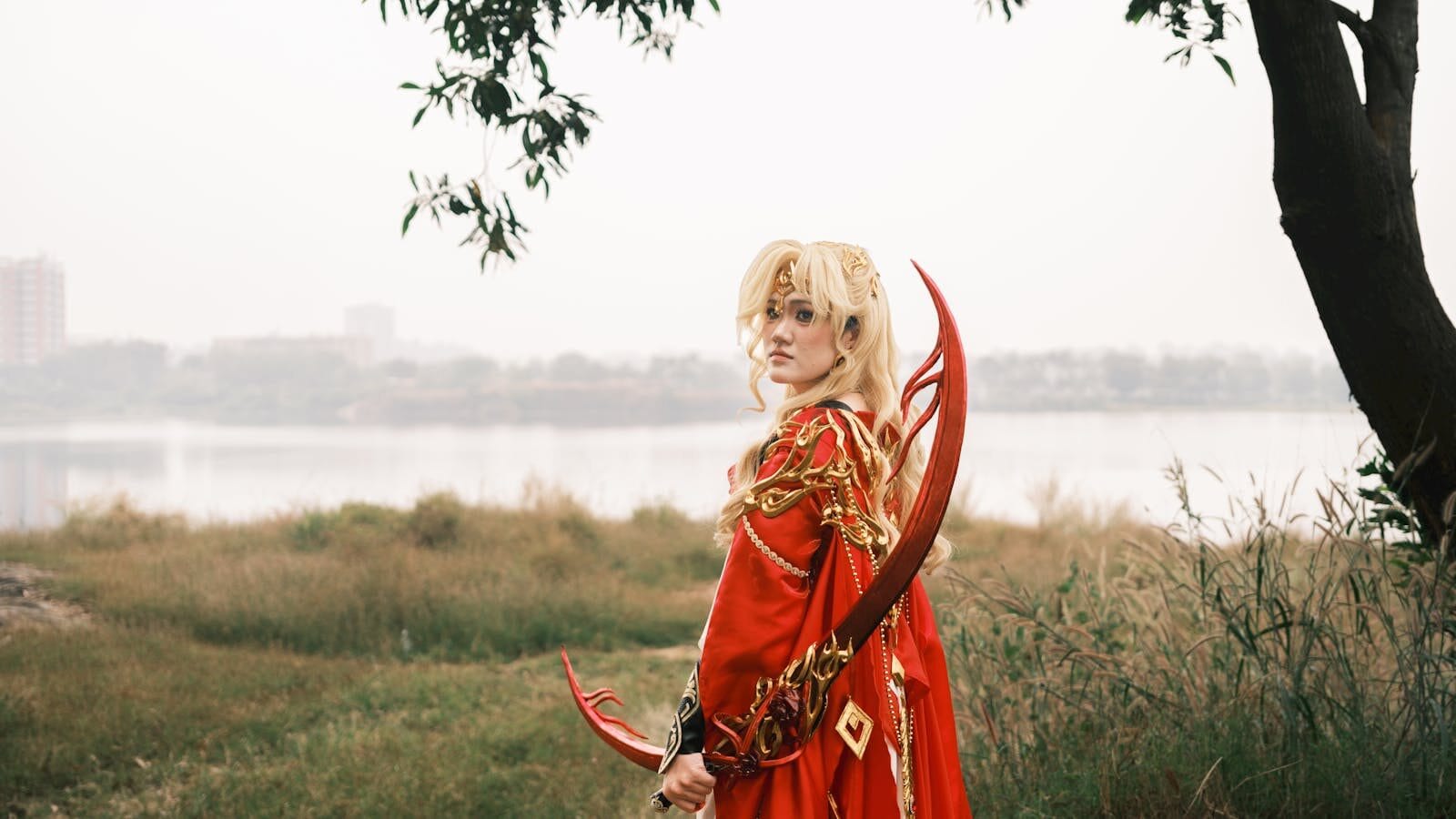 A person in a fantasy cosplay outfit with a red costume stands by a lakeside, holding a decorative bow.